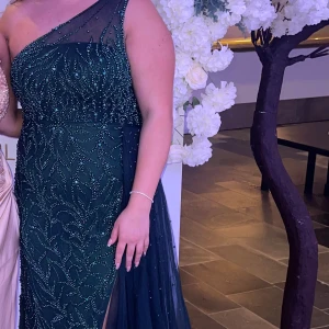 Mörkgrön långklänning med paljetter - Säljer en elegant mörkgrön långklänning med glittriga paljettdetaljer och ett vackert mönster. Klänningen har oneshoulder-design, slits på sidan och ett svepande skirt tyg som ger extra drama och enkelt kan tas bort. Perfekt för bal eller fest! Fråga gärna om fler bilder eller mått.