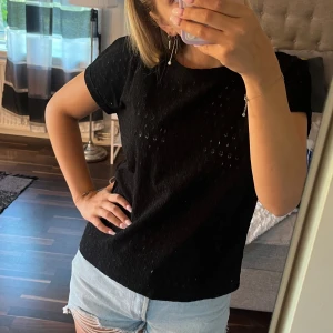 Svart blus från Jyothi med hålmönster - Svart blus från Jyothi i storlek S. Toppen har korta ärmar och är tillverkad i ett  hålmönstrat tyg som ger en luftig känsla. Passformen är normal och toppen är perfekt att styla med jeans eller kjol💙