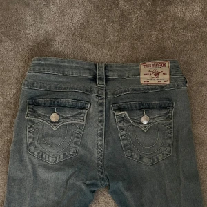 True Religion Joey Low Rise Flare Jeans - Strl 26. Säljer ett par ljusblå True Religion Joey jeans med låg midja och flare-ben. Klassiska stora fickor med silverknappar och broderad logga bak. Jeansen har en snygg tvätt och är gjorda i mjukt denim. Perfekt för dig som gillar retrovibbar och statement-plagg.