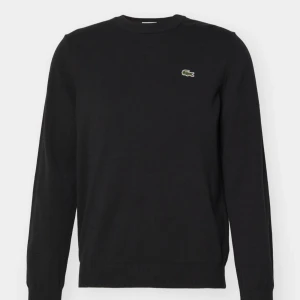 Svart sweatshirt från Lacoste - Svart sweatshirt från Lacoste i klassisk passform, med rund hals och det ikoniska krokodilmärket broderat på bröstet. Tröjan har långa ärmar och är tillverkad i mjuk bomull, perfekt för chill dagar. Enkel och stilren design som funkar till allt.