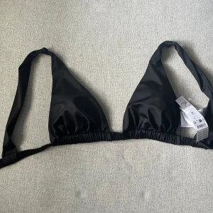 Svart bikinitopp från Gina Tricot - Snygg svart bikiniöverdel från Gina Tricot i storlek XL med breda axelband och klassisk trekantsform. Tillverkad av 83% återvunnen polyester, perfekt för strandhäng. Modellen har justerbar snörning för bästa passform och stilren look. Prislapp kvar