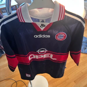 Bayern München retrotröja Adidas - Snygg vintage Bayern München fotbollströja från Adidas med klassisk krage och tryckta loggor. Marinblå bas med röda och vita detaljer, samt breda röda ränder över bröstet och ärmarna. Materialet är lätt och ventilerande polyester, perfekt för match eller samling.