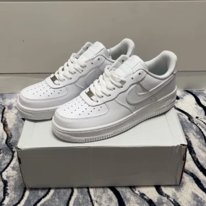 Nike Air Force 1 vita sneakers - Fräscha Nike Air Force 1 sneakers i helvitt med klassisk design och perforerad tå. Skorna har snörning, platt sula och rund tå. Tillverkade i skinn med mjukt foder och tydlig Nike-logga på sidan och hälen. Perfekta för dig som gillar stilrena och ikoniska sneakers.