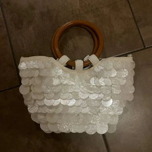 Vit handväska med snäck paljetter - Säljer en unik vit handväska med stora glansiga paljetter på framsidan och snygga runda bruna handtag. Väskan har en cool form och är perfekt för dig som gillar att sticka ut. Materialet är tyg med paljett-detaljer och bruna handtag i plats
