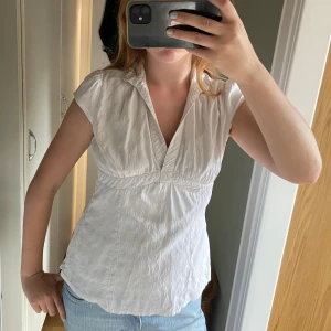 Vit blus med v-ringning och struktur - Säljer en vit blus med v-ringning och korta ärmar. Blusen har ett subtilt strukturerat mönster i tyget och markerad midja för en snygg passform. Perfekt till jeans eller kjol för en fräsch look. Tyget känns lätt och luftigt, perfekt för varmare dagar.