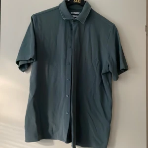 Blå kortärmad skjorta från Zara - Snygg blå skjorta från Zara i regular fit med korta ärmar och klassisk krage. Skjortan har knappar hela vägen framtill och är gjord i mjuk polyester som känns skön mot huden. Perfekt för dig som gillar stilrena och enkla plagg.