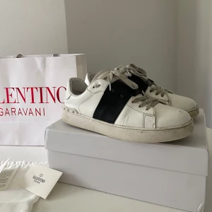 Valentino Garavani Open sneakers vit/svart - Säljer ett par Valentino Garavani Open sneakers i vit skinn med bred svart läderdetalj över sidan. Klassisk rund tå, platt sula och snörning framtill. Ikoniska nitar baktill och medföljer originalpåse, extra skosnören och box. Perfekt för dig som gillar lyxiga street vibes.