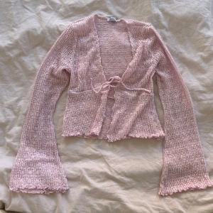 Ljusrosa ribbad omlottkofta med knyt - Superfin ljusrosa kofta med ribbad struktur och omlottmodell. Tröjan har långa ärmar med lätt utsvängda muddar och knyt framtill. Perfekt att styla med jeans eller kjol för en söt look. Använd en gång 