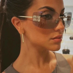 Bruna solglasögon från Miu Miu - Snygga bruna solglasögon från Miu Miu med rektangulär form och guldiga detaljer på skalmarna. Linsen är mörkbrun och skalmarna har Miu Miu-loggan i guld. Kommer med original rosa fodral, tygpåse och certifikat. Perfekt för dig som vill ha en lyxig vibe.