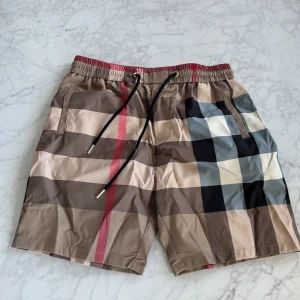 Rutig Burberry shorts med snörning - Snygga badshorts från Burberry med klassiskt rutigt mönster i beige, svart, röd och grå. De har elastisk midja med svart snörning och praktiska fickor. Perfekt för sommaren och ger en lyxig vibe till din outfit.