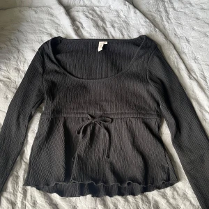 Svart långärmad topp från Nelly - Svart långärmad topp från Nelly med ribbad struktur och rundad urringning. Toppen har en söt knytning i midjan och lätt volangkant nertill. Perfekt för dig som gillar en feminin och trendig stil.