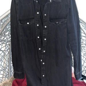 Svart jeansskjorta med knappar - Säljer en svart jeansskjorta med långa ärmar och vita knappar framtill. Skjortan har två bröstfickor med knappstängning och klassisk krage. Perfekt för dig som gillar en cool och avslappnad stil.