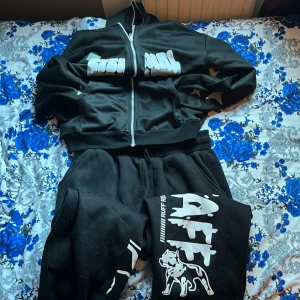 Svarta mjukisbyxor från AISTFF - Svarta mjukisbyxor från AISTFF med stor vit print längs benet och en cool hundgrafik. Byxorna har elastisk midja och är gjorda i mjukt polyester-material. Passformen är loose och de har en avslappnad streetwear-vibe, perfekt för chill eller häng med kompisar. Den kan också passa nån som tar M .