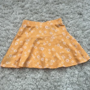 Gul blommig kortkjol - Supersöt gul kortkjol med vita och rosa blommor. Kjolen har en lätt utsvängd form och är gjord i mjukt bomullstyg som känns skönt mot huden. Perfekt för soliga dagar och ger en riktigt glad vibe till din outfit.