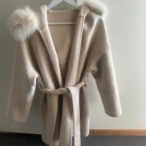 Beige kappa med päls - Snygg beige ull kappa med stor huva kantad av fluffig ljus äkta päls. Jackan har bälte i midjan för en skön passform och är tillverkad i ett mjukt ull material. Perfekt för dig som vill ha en stilren och mysig look under kyliga dagar. Storlek xs 