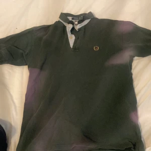 Grön pikétröja Tommy Hilfiger  - Säljer en grön pikétröja från Tommy Hilfiger i storlek L fast den är för liten för mig som är 170 cm. Tröjan har klassisk krage med randigt tyg på insidan och knappar framtill. På bröstet finns ett broderat emblem med lejon och lagerkrans. Materialet är mjuk bomull och modellen har korta ärmar.