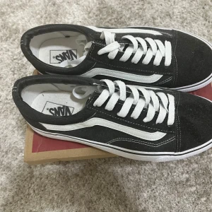 Svarta Vans Old Skool sneakers - Svarta Vans Old Skool sneakers med klassisk vit sidorand och vit snörning. Skorna har en låg profil, rund tå och platt gummisula med det ikoniska rutmönstret undertill. Ovandelen är i mocka och canvas, vilket ger en skön och cool vibe.