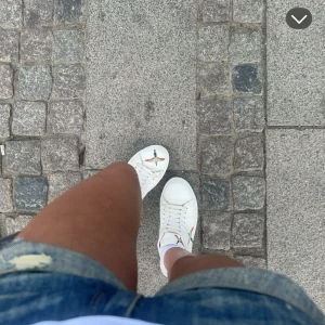 Axel Arigato skor - Säljer ett par vita sneakers med rund tå och platt sula. Skorna har snörning och är dekorerade med färgglada fågelmotiv på sidorna och ovansidan. Kom med pris förslag 💗