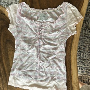 Rutig blus med snörning från ZE-ZE - Supersöt rutig blus från ZE-ZE i ljusrosa och vit med diskreta blå linjer. Blusen har korta puffärmar, snörning framtill och en söt volangkant vid halsen. Materialet känns lätt och luftigt, perfekt för varmare dagar. Passformen är normal och toppen har en feminin vibe.