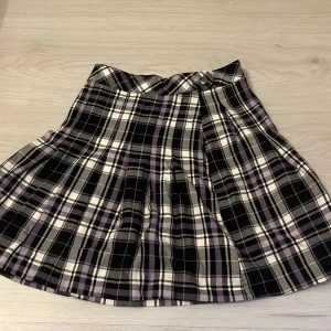 Rutig kortkjol från Lindex i lila och svart - Superfin rutig kortkjol från Lindex i svart, vitt och lila. Kjolen har klassiskt rutmönster, knäppning fram och är lätt utställd. Perfekt för dig som gillar en preppy vibe och vill sticka ut lite extra med färg. Passar till både sneakers och boots.