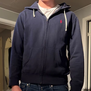 Marinblå hoodie från Ralph Lauren - Säljer en marinblå hoodie från Ralph Lauren med dragkedja och huva med vita snören. Tröjan har det klassiska röda Polo-logot på bröstet, två fickor framtill och är långärmad. Perfekt för chill dagar och enkel att matcha med jeans. Storlek s hyfsat använd 