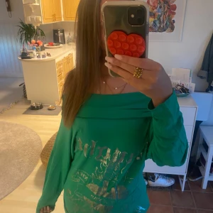 Grön offshoulder tröja Seventy Seven - Cool grön långärmad tröja från Seventy Seven med offshoulder-design och metallictryck med texten 'Naughty' och motiv på framsidan. Tröjan är i mjuk bomull och har en loose passform som sitter skönt och avslappnat. Perfekt för dig som gillar statement-plagg! (Aldrig använd)