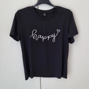 Svart t-shirt med happy-tryck  - Svart t-shirt med vit text 'happy' och ett litet hjärta på bröstet. T-shirten har en klassisk rund hals och korta ärmar med mjuk och skön bomullskvalitet. Enkel och stilren design som ger en positiv vibe. Från Shein i storlek M 💓💓