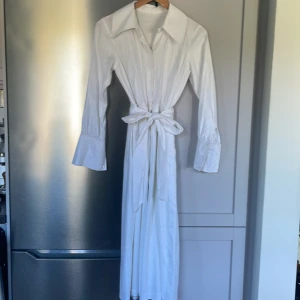 Vit långklänning med knyt i midjan - Snygg vit långklänning med bred krage och långa ärmar med breda manschetter. Klänningen har knytband i midjan för en markerad siluett och är tillverkad i ett lätt, glansigt material som ger en elegant känsla. Perfekt för dig som gillar klassisk och stilren design. Klänningen har lite märken som ni ser på bilden. Det var skräddarsydd. Jag är 1m60 och 49kg