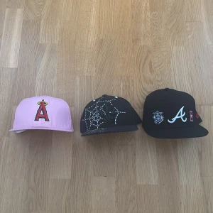 NewEra Kepsar - Säljer tre kepsar: en ljusrosa med röd och gul broderad 'A' framtill, en svart med vit spindelnätsmönster och broderad spindel, samt en svart med vit 'A' och flera broderade märken på sidan. Perfekt för dig som vill sticka ut med coola detaljer och färger.