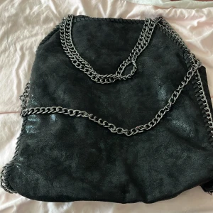 Svart axelväska med kedjedetaljer - Snygg svart axelväska med grova silverfärgade kedjor som axelrem och dekor längs kanterna. Väskan har en mjuk mockaliknande yta och en stilren, fyrkantig form. Perfekt för dig som gillar en edgy look och vill ha plats för allt viktigt.