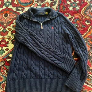 Marinblå stickad tröja Polo Ralph Lauren - Säljer en marinblå stickad tröja från Polo Ralph Lauren med half zip och kabelstickat mönster. Tröjan har långärmade ribbade muddar, hög krage och röd broderad logga på bröstet. Perfekt för dig som gillar klassisk och chill stil och perfekt till hösten.