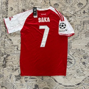 Arsenal Saka #7 Champions League tröja - Riktigt snygg Arsenal fotbollströja från Adidas med Saka och nummer 7 på ryggen. Röd kropp med vita korta ärmar, vita detaljer och Champions League-märke på ärmen. Materialet är lätt och ventilerande polyester, perfekt för match eller träning.
