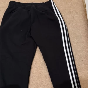 Svarta Adidas mjukisbyxor med ränder - Svarta mjukisbyxor från Adidas med klassiska vita ränder längs sidorna och Adidas-logga på låret. Byxorna har elastisk midja med snörning och är gjorda i mjukt bomullsmaterial. Perfekta för chill eller träning, riktigt sköna och stilrena.