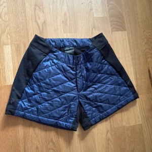 Quiltade blå shorts Peak Performance - Storlek: XS Snygga quiltade shorts från Peak Performance i marinblått med svarta paneler. De har en loose passform, dragkedja och knapp framtill. Materialet är polyester och fleece, vilket gör dem perfekta för träning eller utomhusaktiviteter under kyligare dagar.