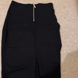 Svart pennkjol med dragkedja  - Snygg svart pennkjol i storlek S. Kjolen har en guldig dragkedja bak och är figurnära med rak siluett. Perfekt för dig som gillar stilrena och enkla plagg. Materialet är mjukt och stretchigt, vilket gör den bekväm att bära.