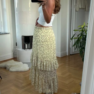 Blommig volangkjol i ljusgult - Supersöt långkjol i ljusgul ton med blommigt mönster och volanger. Kjolen har en luftig och flowy känsla, perfekt för varma dagar. Resår i midjan gör den bekväm och enkel att bära. Materialet är tunt och lätt.