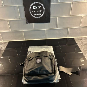 Svart axelväska från Stone Island - Helt ny Snygg svart axelväska från Stone Island med klassisk kompasslogga på framsidan. Väskan har en fyrkantig form, praktisk framficka med knappstängning och robust dragkedja. Tillverkad i slitstark polyester som är vattentät med justerbar rem för enkel användning.