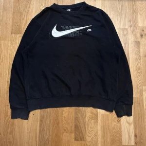 Svart Nike sweatshirt med logga - Svart sweatshirt från Nike i storlek M med stor vit Nike Swoosh och text på bröstet. Tröjan har rund hals, ribbade muddar vid ärmslut och nederkant samt lång ärm. Materialet är mjukt och känns som bomull med insida i fleece. Perfekt för chill eller streetwear.