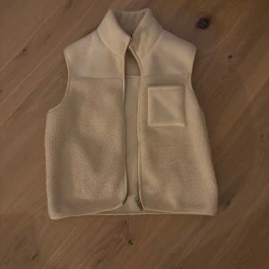 Beige fleeceväst med dragkedja - Säljer en mysig beige fleeceväst med dragkedja och en praktisk bröstficka. Perfekt för lager på lager under kyliga dagar. Västen har en enkel och stilren design. Har knappt använt den, men är så fin!
