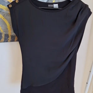 Svart topp från Bodyflirt - Snygg svart topp från Bodyflirt med dekorativa knappar på axlarna. Toppen har en elegant drapering och är ärmlös, vilket gör den perfekt för varmare dagar eller lager-på-lager-stil.