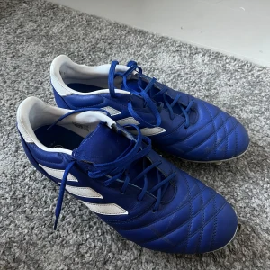 Blå fotbollsskor med snörning från adidas  - Säljer ett par blå fotbollsskor med vita detaljer och snörning från adidas. Skorna har en rund tå och är perfekta för fotbollsträning eller match. De har en klassisk design med dobbar för bra grepp på planen. Storlek 42 2/3. 