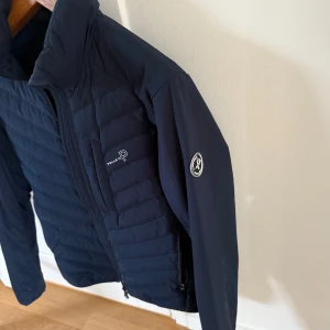 Mörkblå jacka från Pelle P - Snygg mörkblå jacka från Pelle P med dragkedja och broderad logga på bröstet. Jackan har en quiltad design och långa ärmar. Perfekt för kyligare dagar.