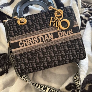 Svart och beige axelväska från Christian Dior - Snygg axelväska från Christian Dior i svart och beige med märkets logotyp. Väskan har ett mönster och detaljer i guld samt en justerbar axelrem. Väldigt lyxig och fin. Använd 3-8ggr sparsamt.