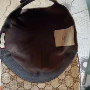 Beige mönstrad keps från Gucci - Snygg beige keps från Gucci med det klassiska GG-mönstret. Kepsen har en justerbar rem baktill och en randig detalj i grönt och rött på sidan. Perfekt för att ge din outfit en lyxig touch.