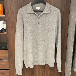 Stickad Pikétröja Grå Davida - Säljer en elegant grå kashmirtröja från Davida. Tröjan har en klassisk krage och knappar framtill. Perfekt för en stilren look. Den är långärmad och tillverkad i 100% kashmir. Den har inga defekter och är behandlad väl. Superfin!