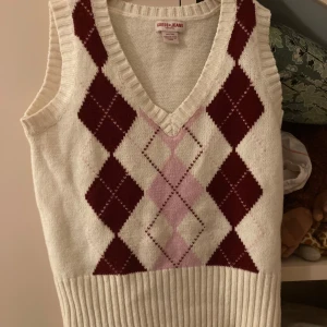 Vit stickad väst från Guess Jeans - Säljer en snygg vit stickad väst från Guess Jeans med ett klassiskt rutmönster i vinrött och rosa. Västen har en v-ringad design och ribbade kanter. Använd 1 gång. Köpt på beyond retro. Storlek är XL men skulle säga att den sitter mer som S/M.