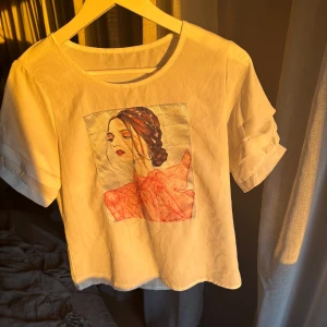 Vit t-shirt med tryck  - Säljer en vit t-shirt med ett konstnärligt tryck av en kvinna. T-shirten har korta ärmar med en liten volangdetalj. 