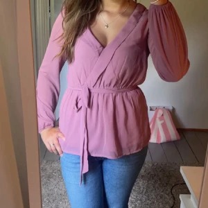 Rosa blus med knytband - Söt rosa blus med knytband i midjan. Blusen har långa ärmar och en v-ringad desig. Perfekt för både vardag och festliga tillfällen.