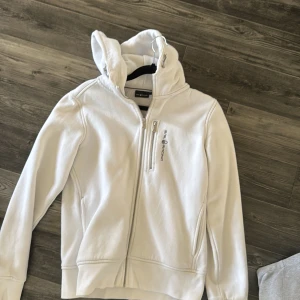 Vit jacka från Sail Racing - Säljer en vit jacka från Sail Racing med dragkedja och huva. Jackan har en bröstficka med dragkedja och logotyp på bröstet. Perfekt för en stilren och sportig look.