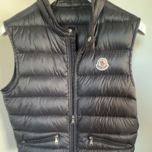 Svart gui väst från Moncler - Snygg svart gui väst från Moncler med dragkedja och logotyp på bröstet, strl xxs. Perfekt för varmare dagar nu inför våren/sommaren. Västen har praktiska fickor med dragkedja och en stilren look. Den är knappt använd och är i mycket bra skick 9/10. Det finns kvitto kvar från Harrods, ny pris var 300 pund. Allt annat finns också kvar och de medföljer extra knappar i fall nån går sönder och traveling case, som ni ser på bilderna. Kontakta mig om det finns nån fråga!